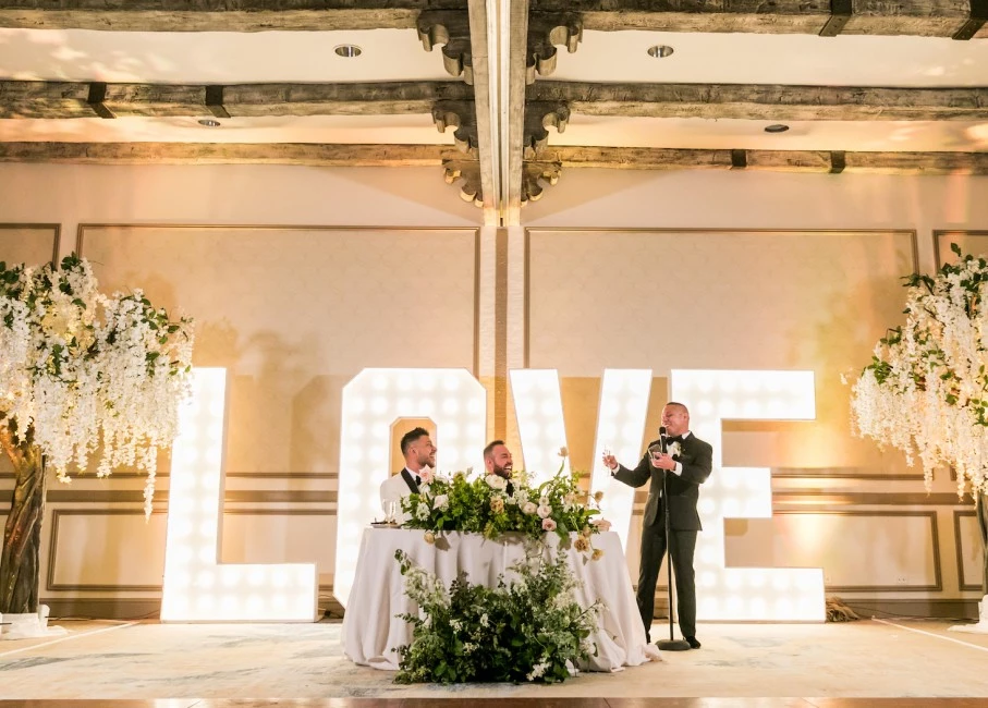 a man speaking into a microphone at a table with flowers and letters