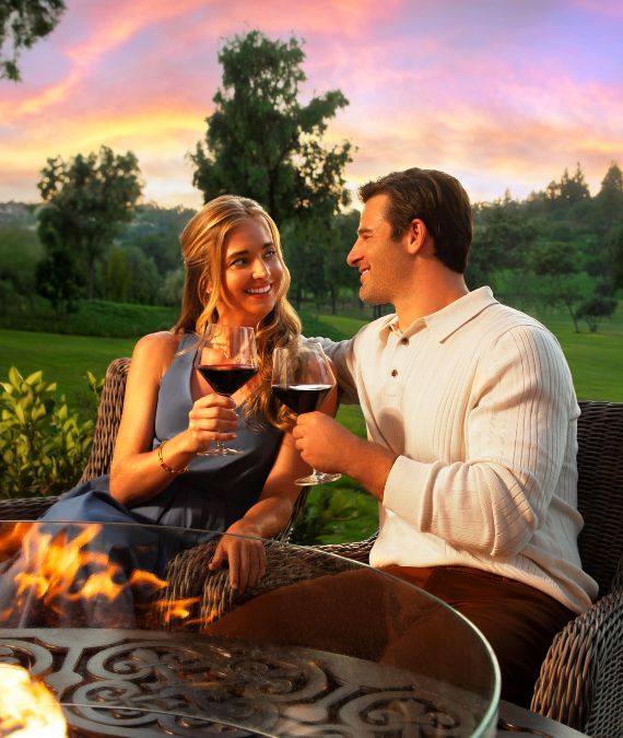 a man and woman holding wine glasses and sitting at a fire pit