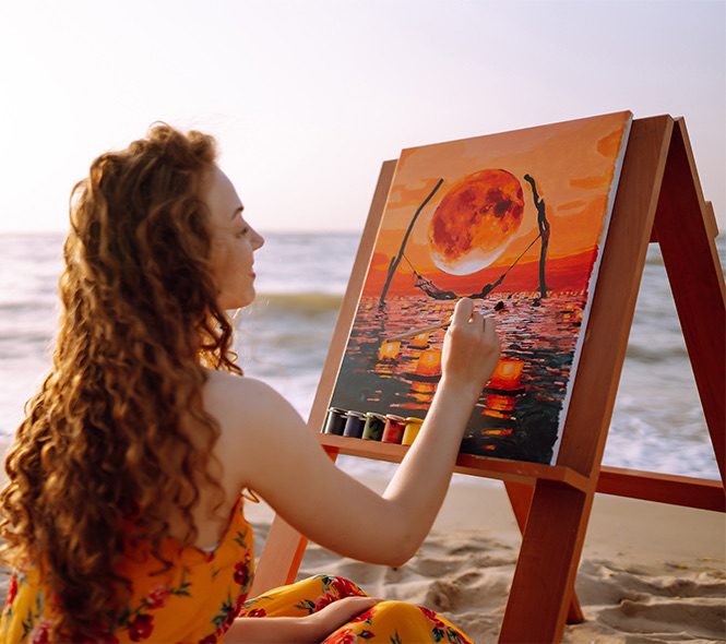 a woman painting on an easel while sitting in the sand