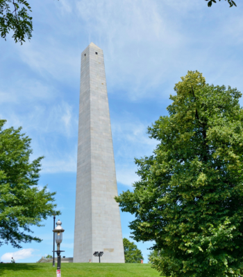 a tall tower with trees in the background with Bunker Hill Monument in the background