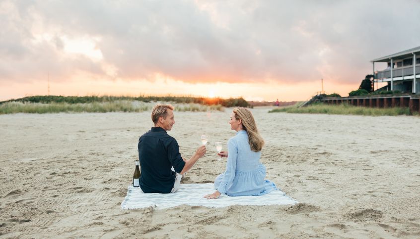 a man and woman sitting on a blanket on a beach with wine glasses