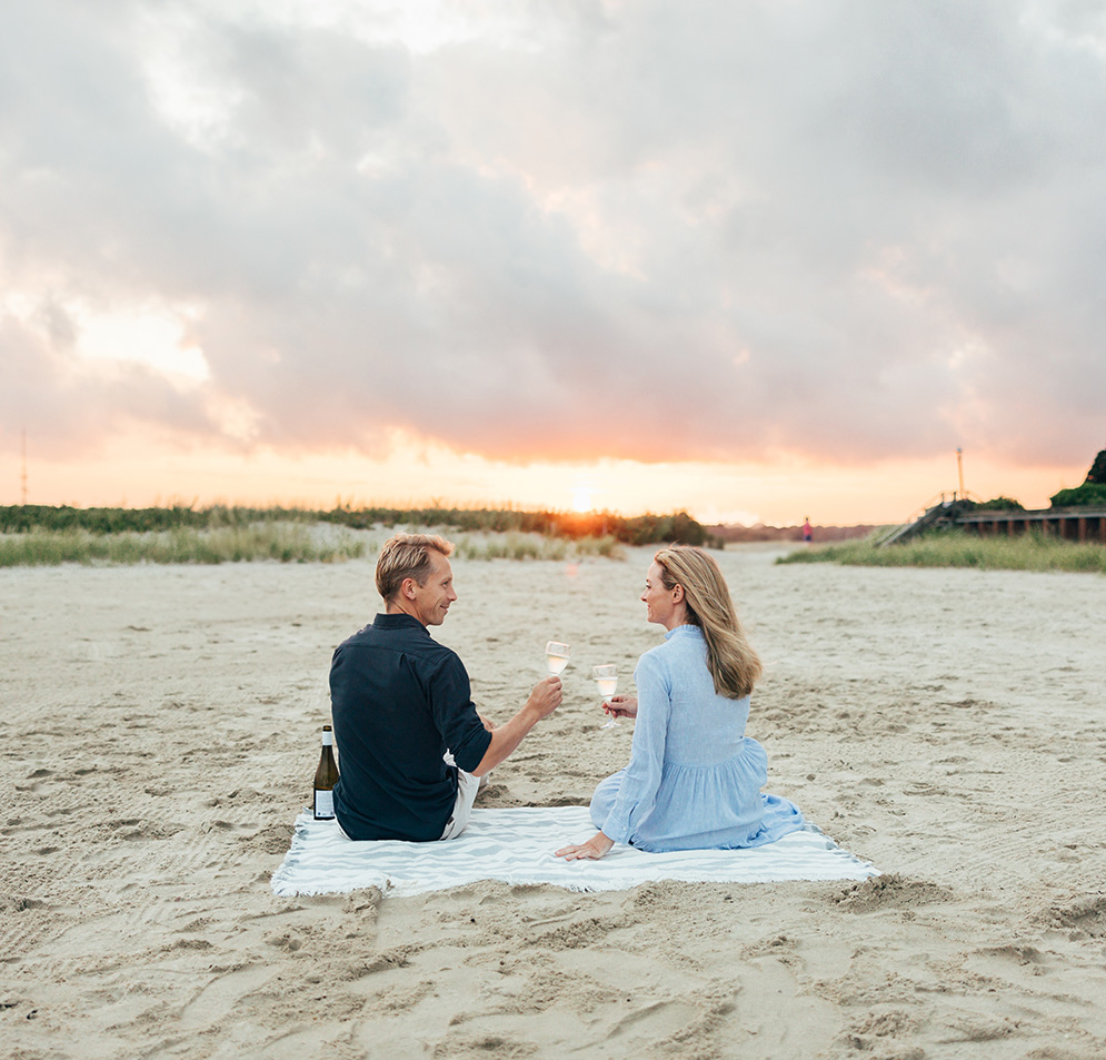 a man and woman sitting on a blanket on a beach with wine glasses