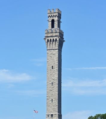 a tall stone tower with a flag with Pilgrim Monument in the background