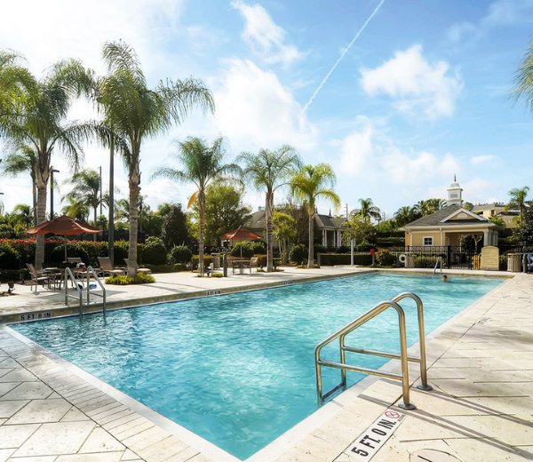 A large outdoor swimming pool with a pool deck, lounge chairs, and palm trees under a blue sky with clouds.