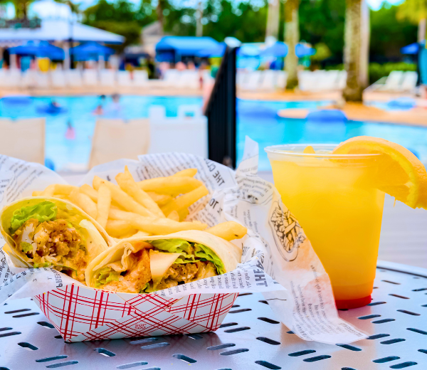 a basket of food and a drink on a table by the pool