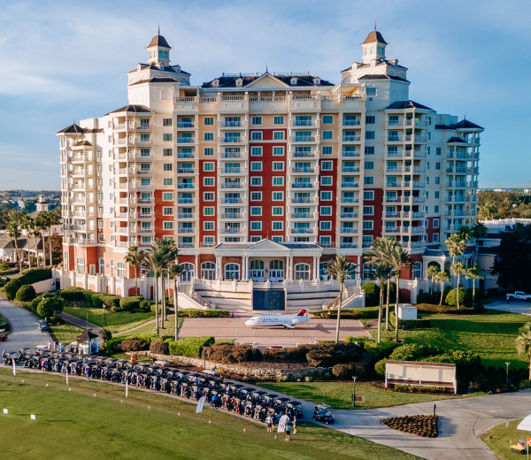 A large hotel building with balconies and towers, surrounded by lawn and palm trees, with a golf course in the foreground.