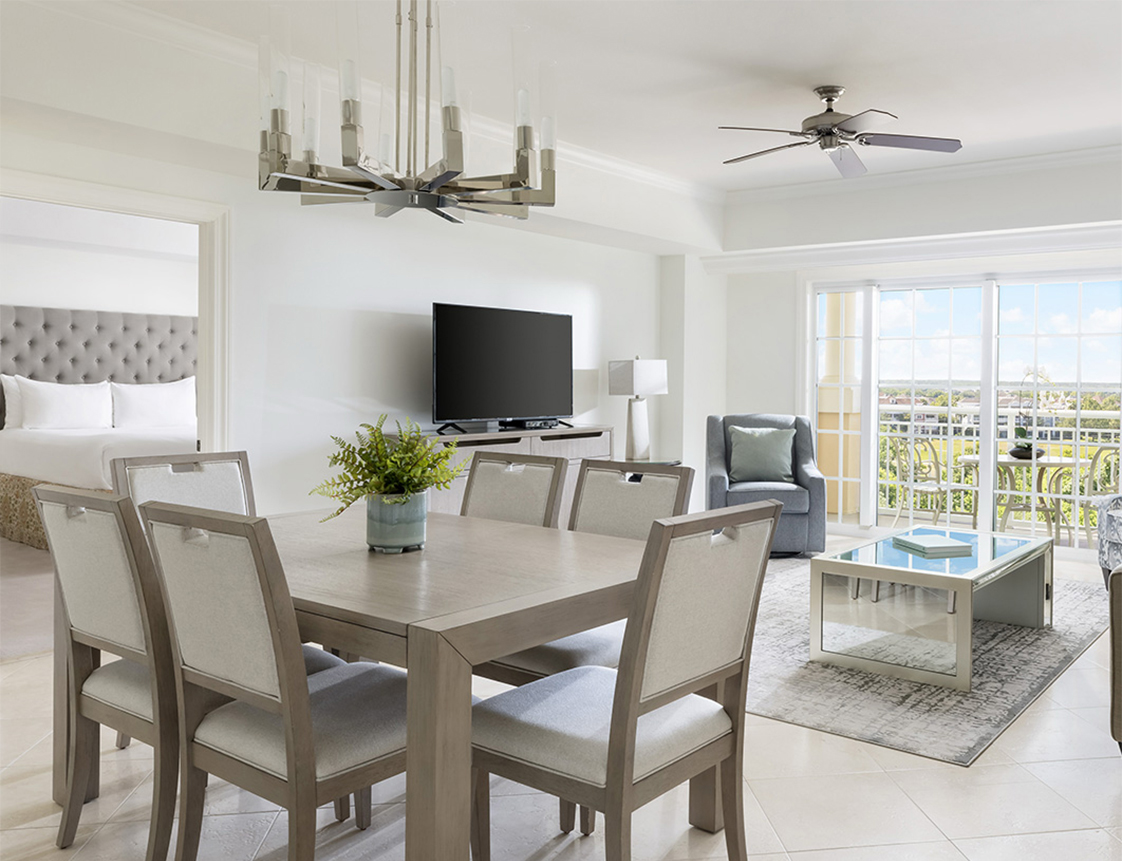 A wide view of a bright, open-plan living space with a dining area, a sitting area, and a partial view of a bedroom.