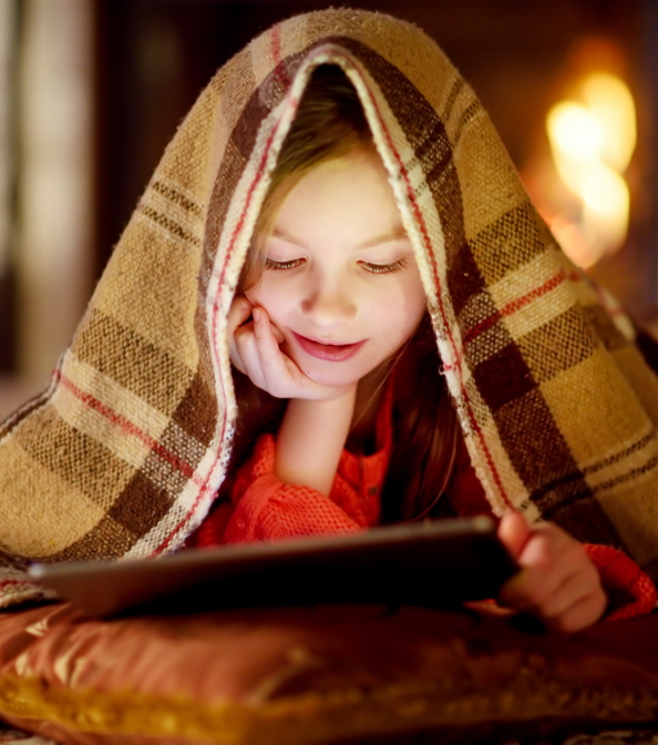 a girl lying under a blanket looking at a tablet