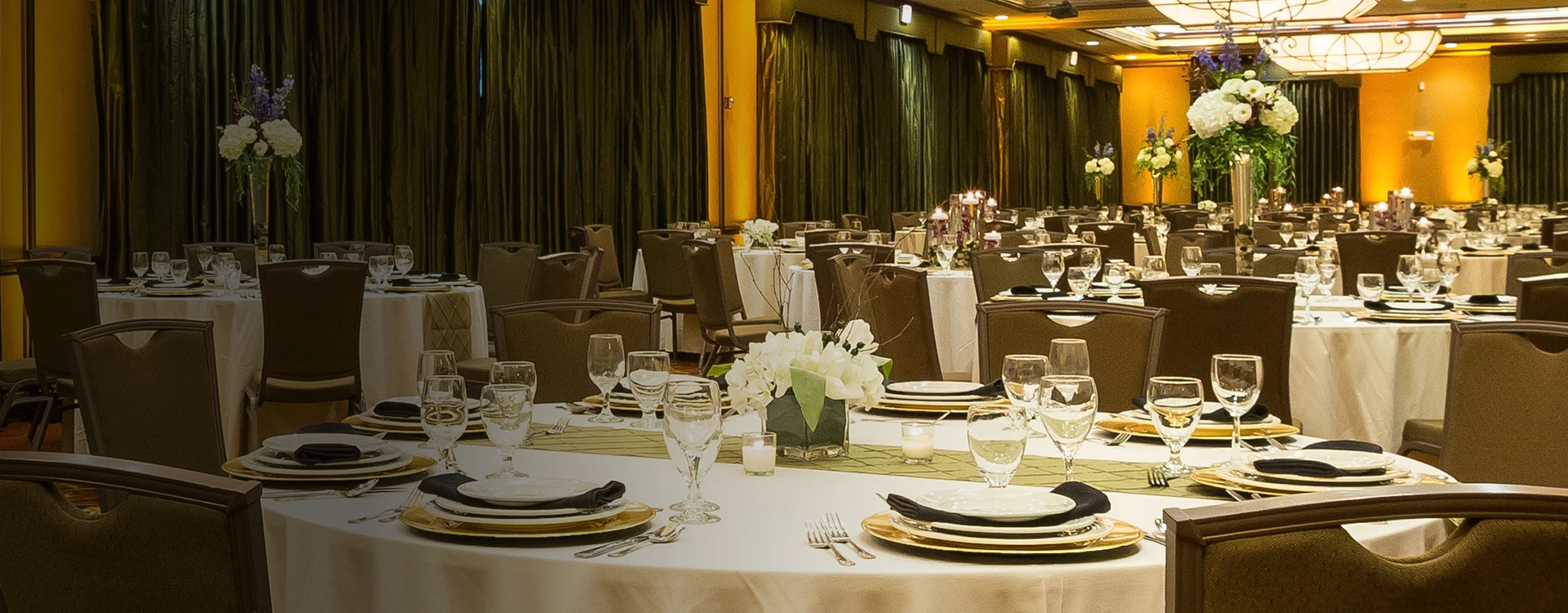 A formal dining room with round tables set for a large event, featuring white tablecloths, floral centerpieces, and candlelight.