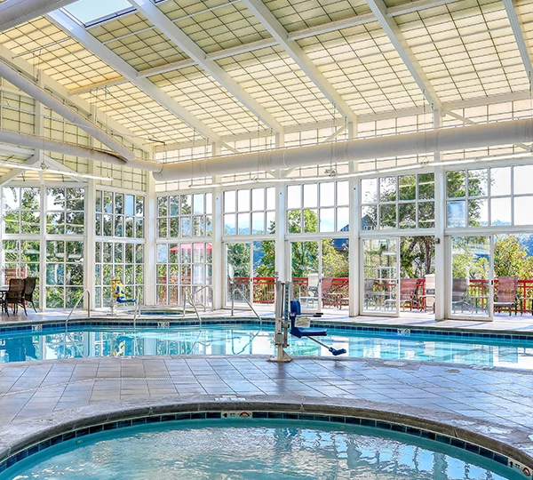 An indoor pool with a curved edge and a tiled floor, surrounded by large windows and a white ceiling with beams.