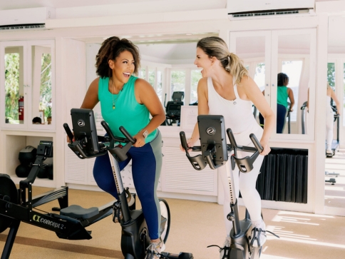 two women in a fitness center smiling at each other while riding exercise bikes