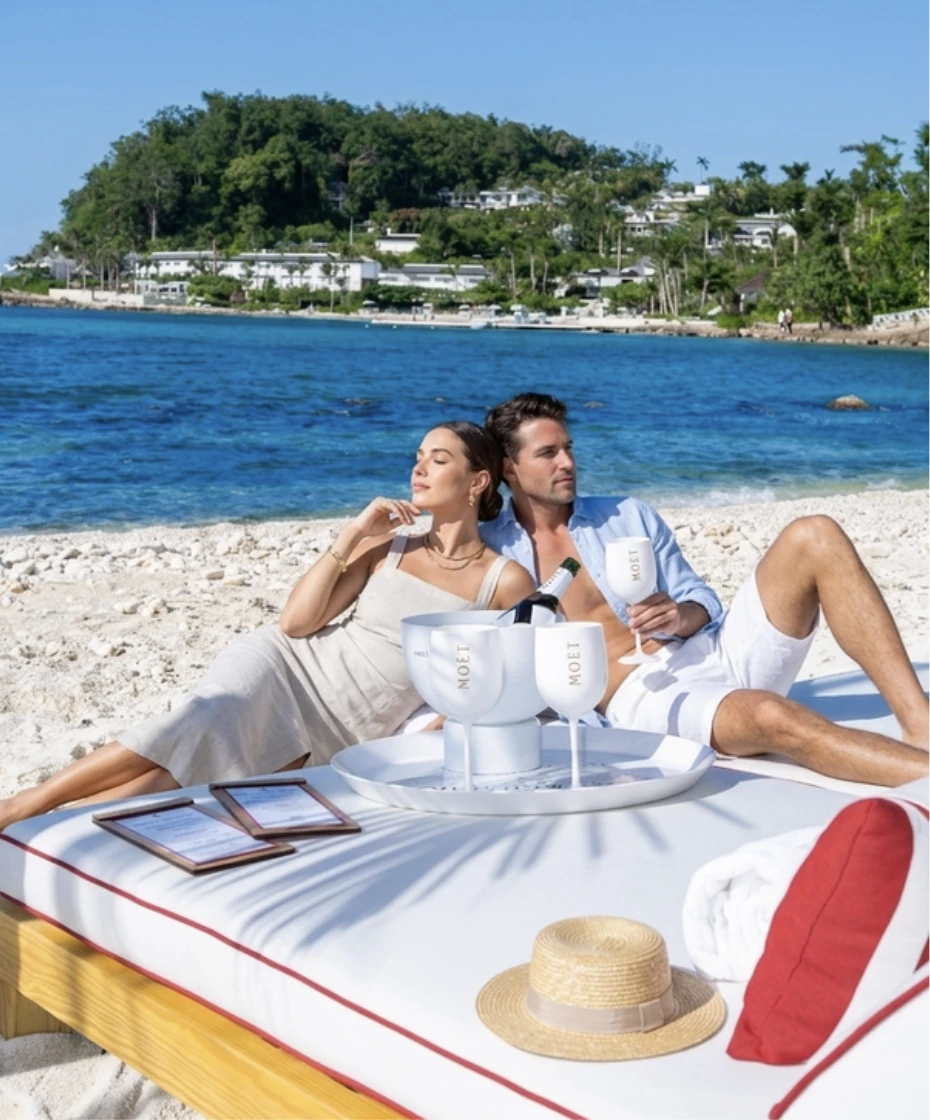 a man and woman sitting on a beach with a table with wine glasses and a body of water