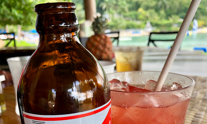 a brown bottle next to a glass of liquid with ice and a straw