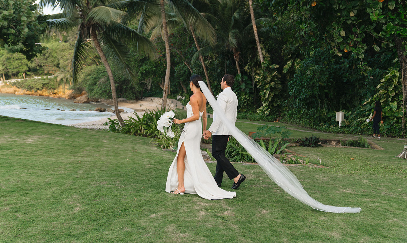 a man and woman walking on grass with palm trees and water