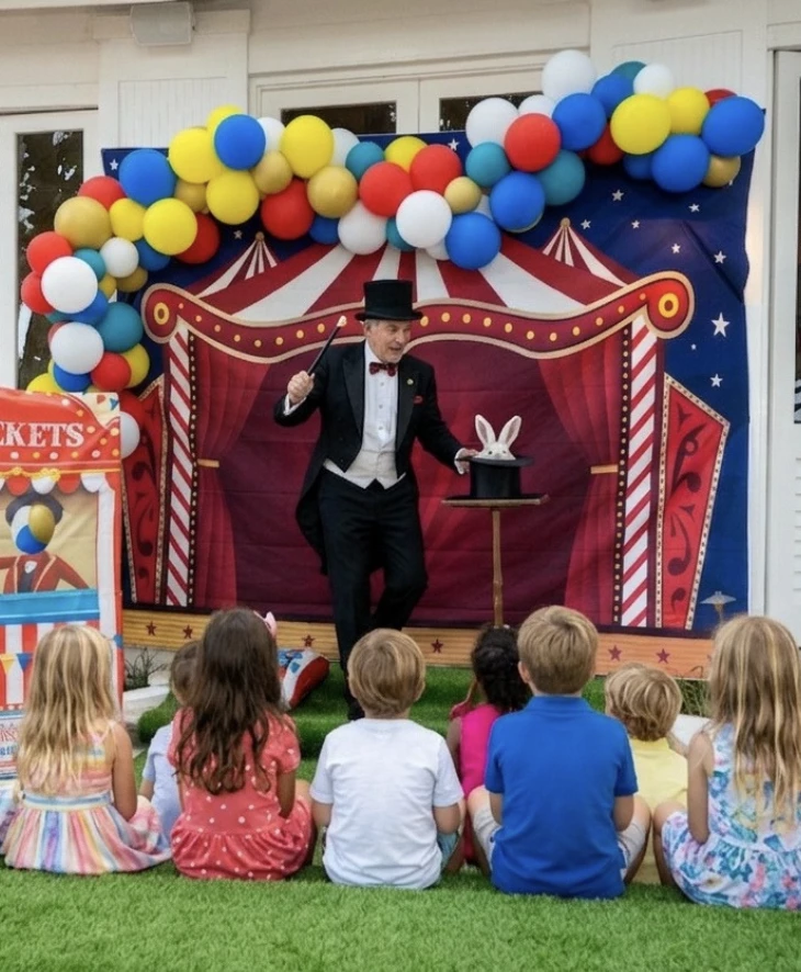 a man in a suit and hat standing on a stage with a group of kids sitting on grass