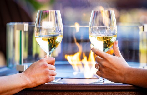 two hands holding wine glasses with yellow liquid in front of a fire