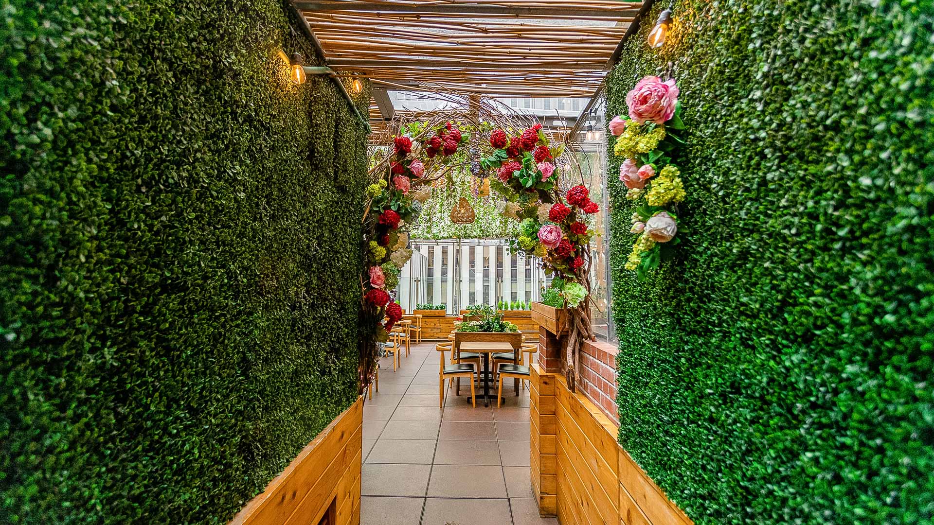 A narrow outdoor dining area with a wooden pergola, surrounded by hedge walls and decorated with a large flower arrangement.
