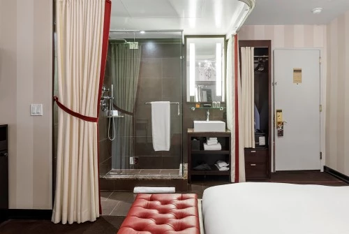 A wide view of a hotel room with a red leather bench, a white bed, and a glass-enclosed shower