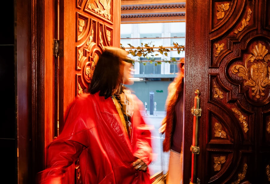 A woman in a red coat walking through an ornate doorway with carved wood and gold hardware.