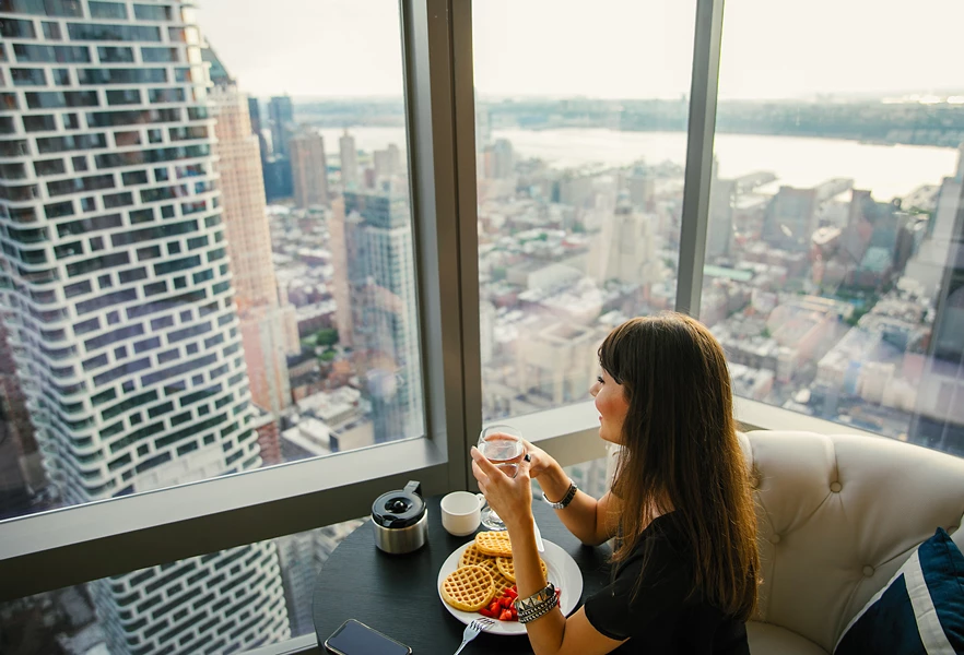 A woman sitting at a table in a room with a city view, eating waffles and drinking from a glass.