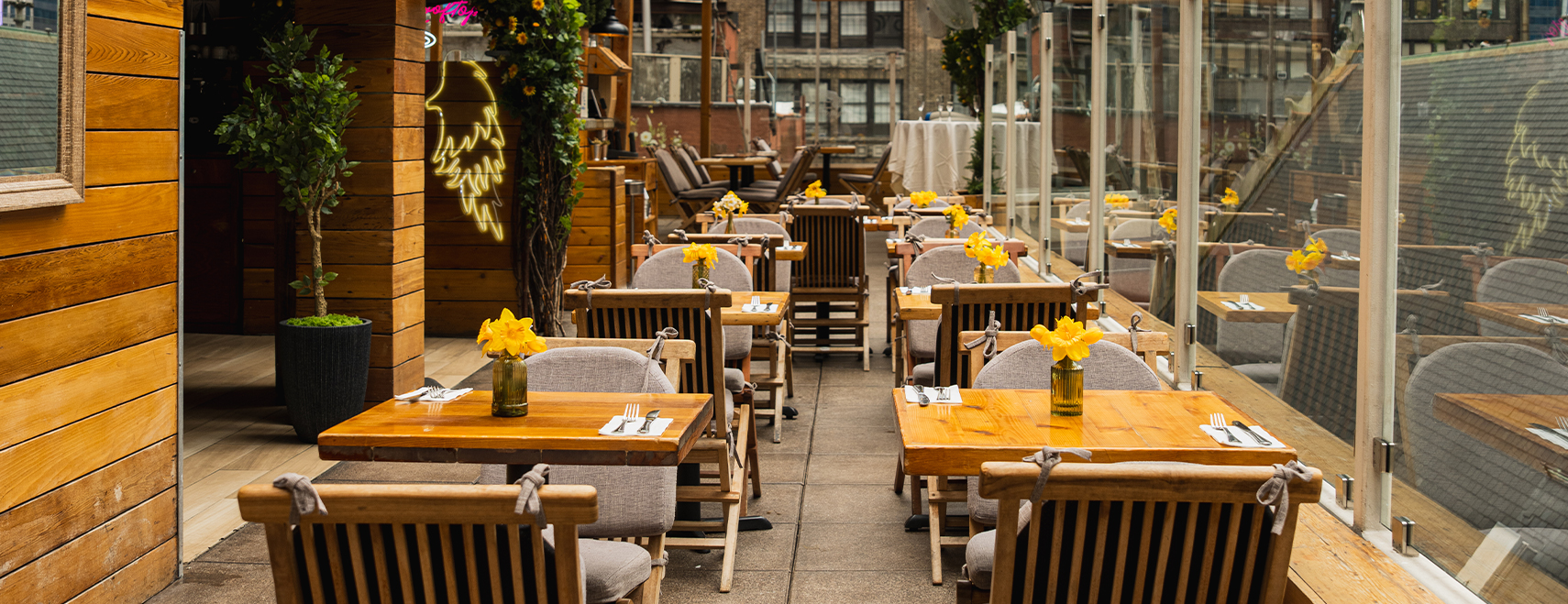 A restaurant patio with wooden tables and chairs, yellow flowers, and glass walls.
