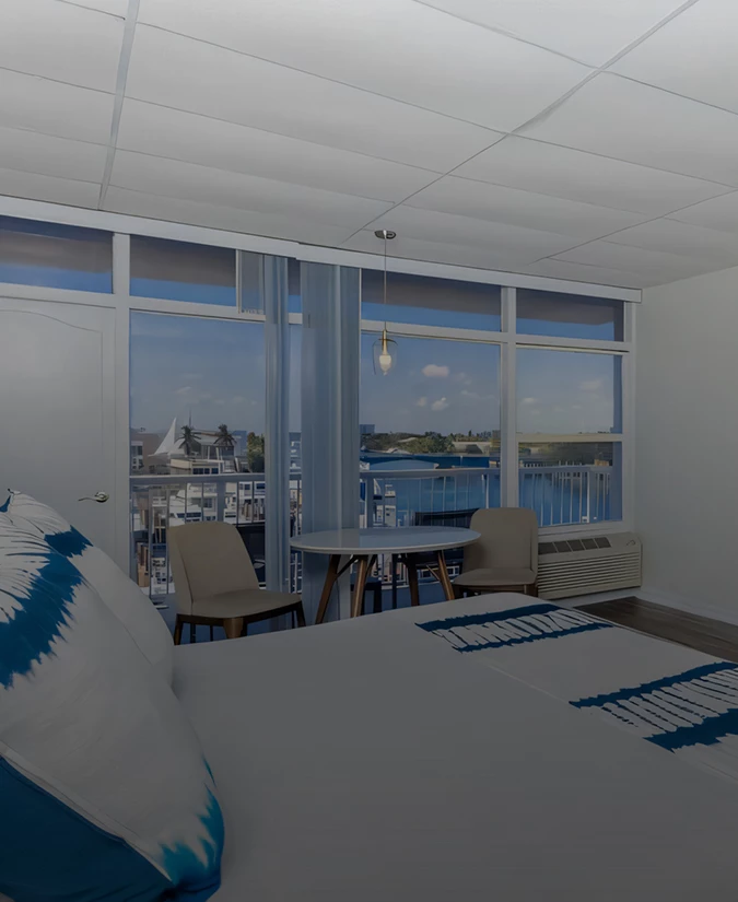 A hotel room with a bed, table, chairs, and a large window with a view of the marina.