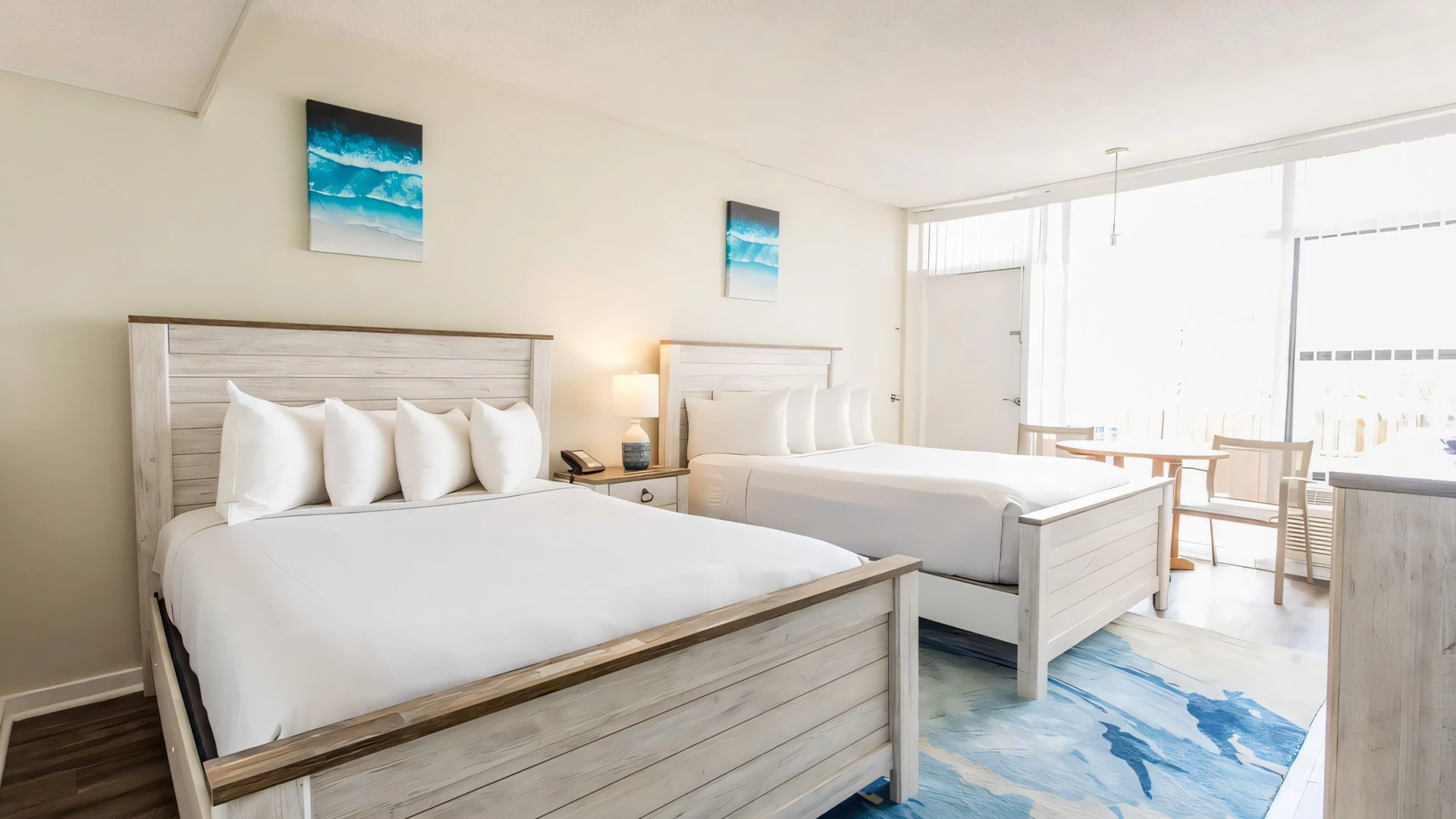 A bright and airy hotel room with two queen beds, a nightstand, and a table with chairs by the window.