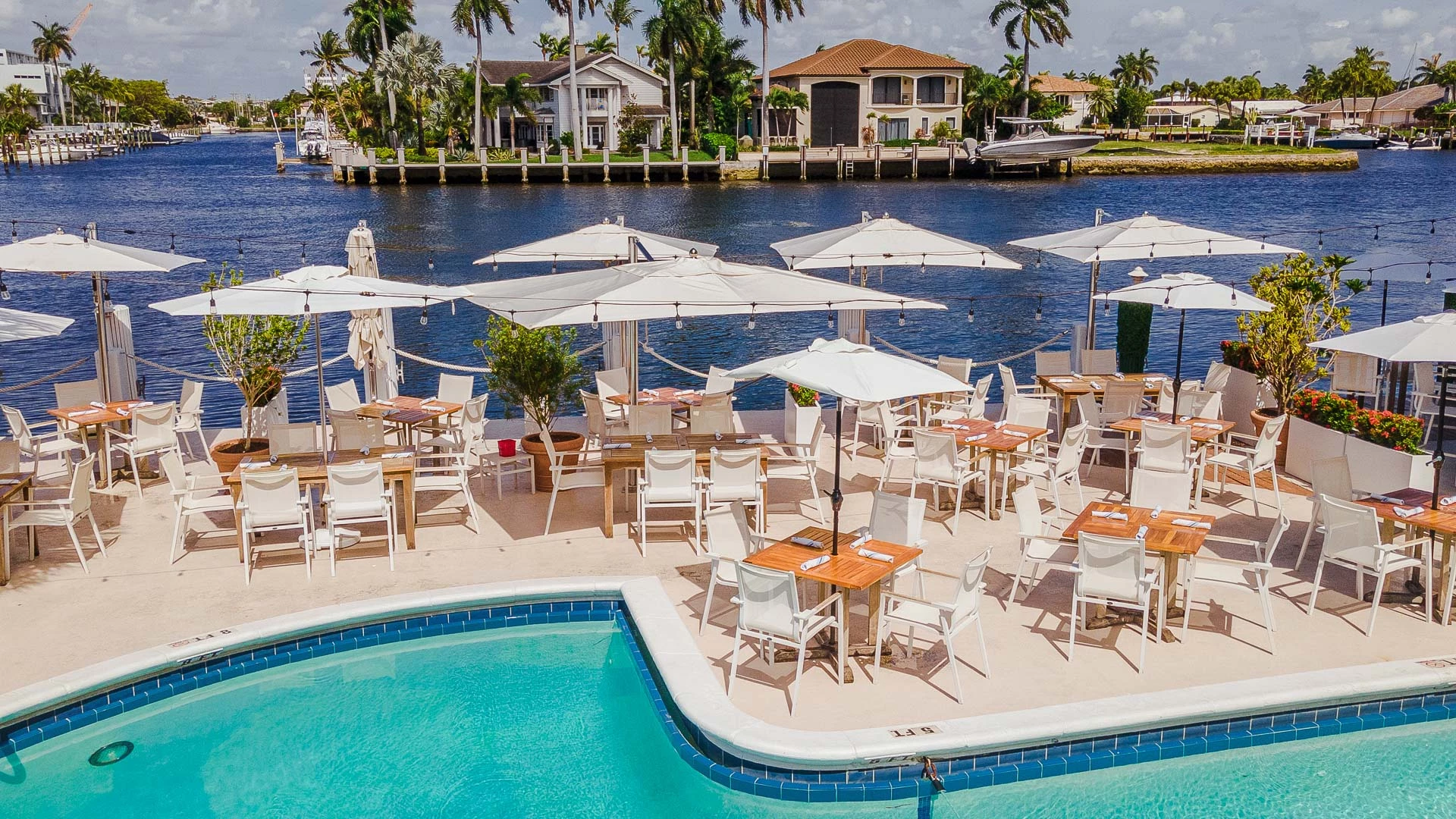 A poolside dining area with white furniture and umbrellas overlooking a body of water.