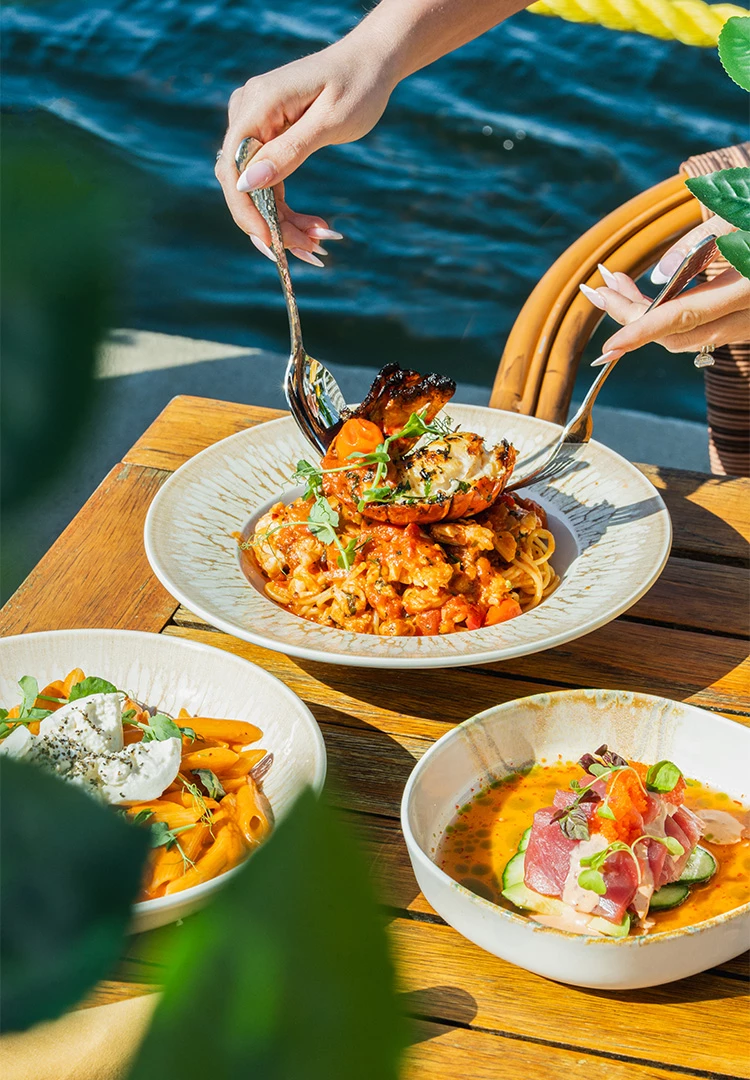 A table with three plates of food, a hand holding a fork and spoon, and a body of water in the background.