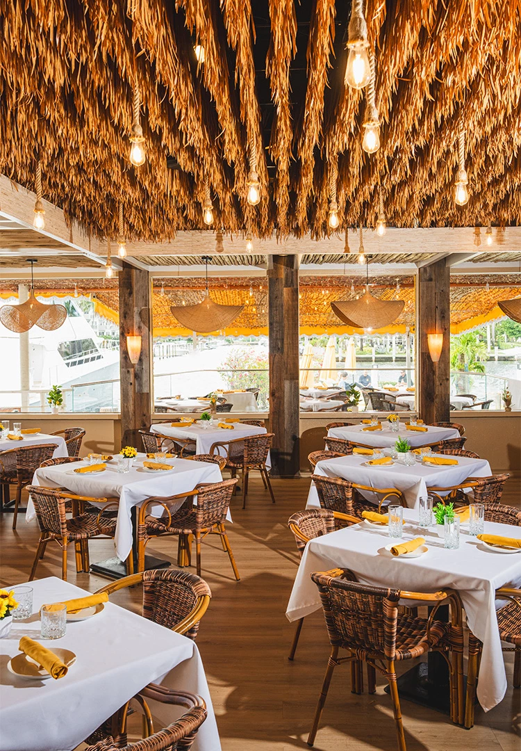 A dining area with white tables, wicker chairs, and a thatched ceiling, overlooking a marina.