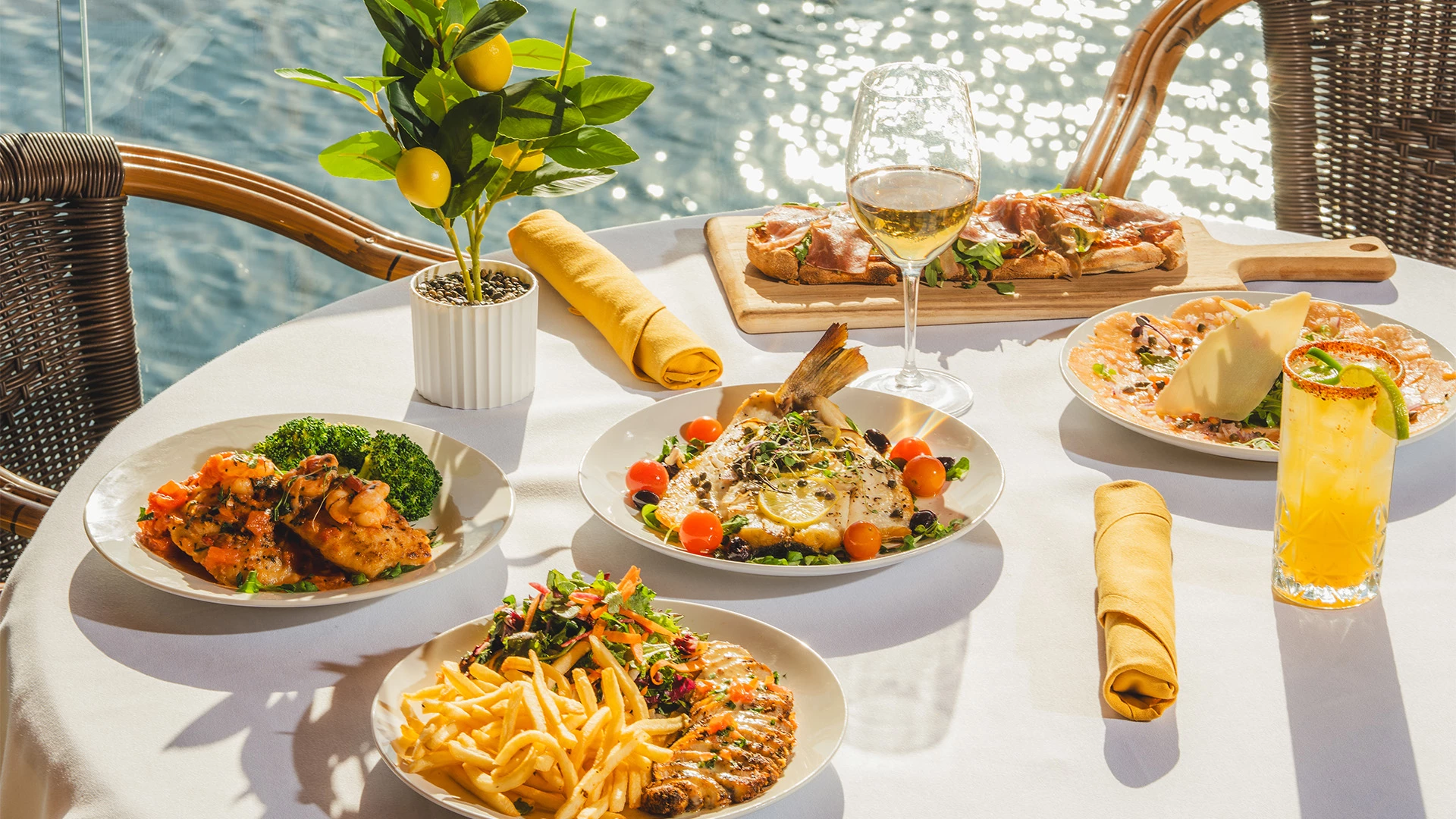 A table set with various dishes and drinks on an outdoor patio overlooking the water.