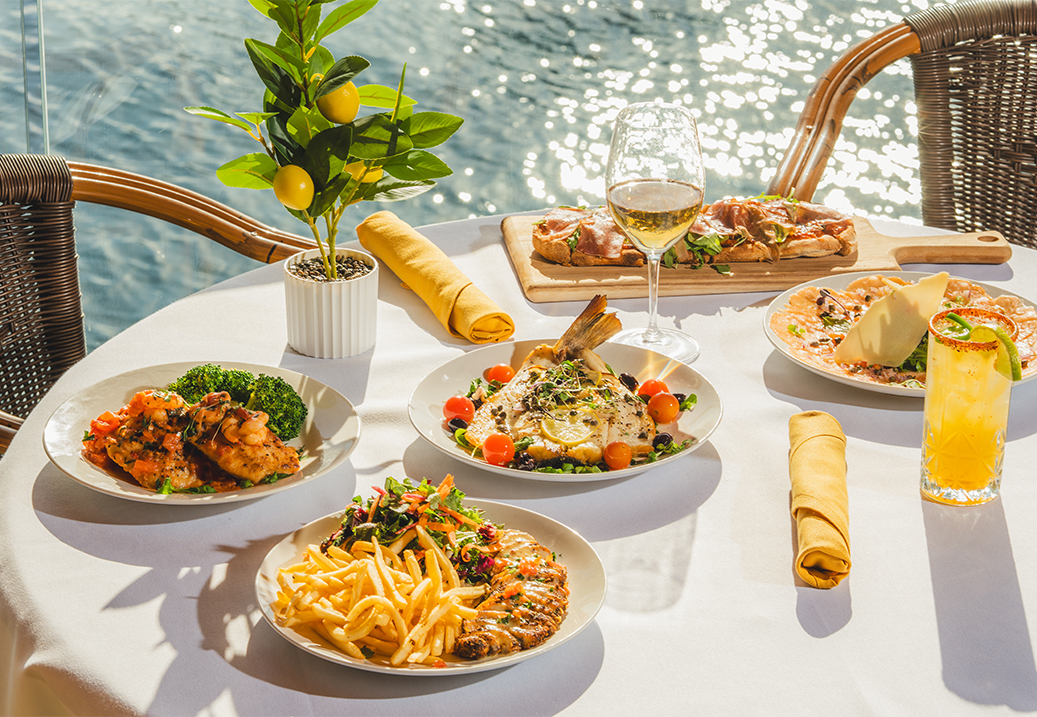a table set with several plates of food and glasses of drinks on a white tablecloth with a water view