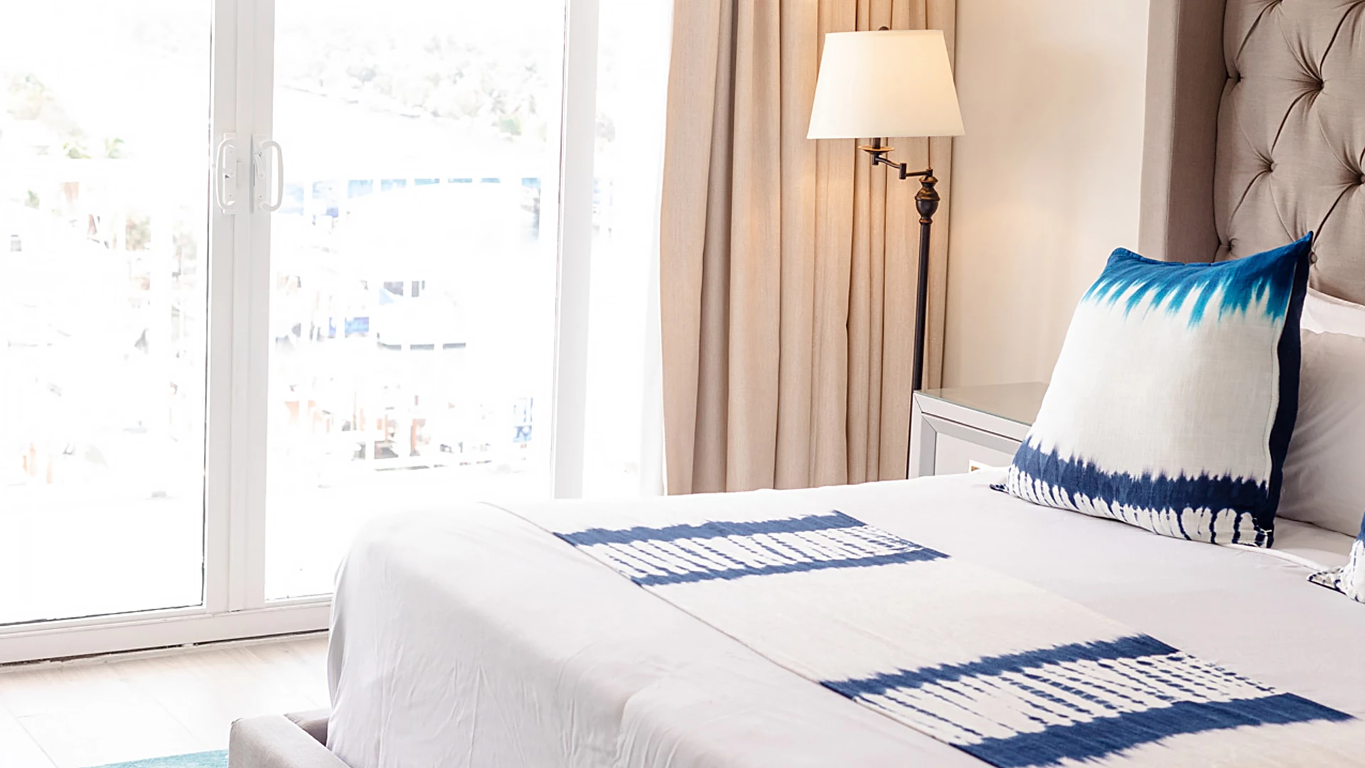 A bed with white sheets and blue accents in a room with a window and lamp.