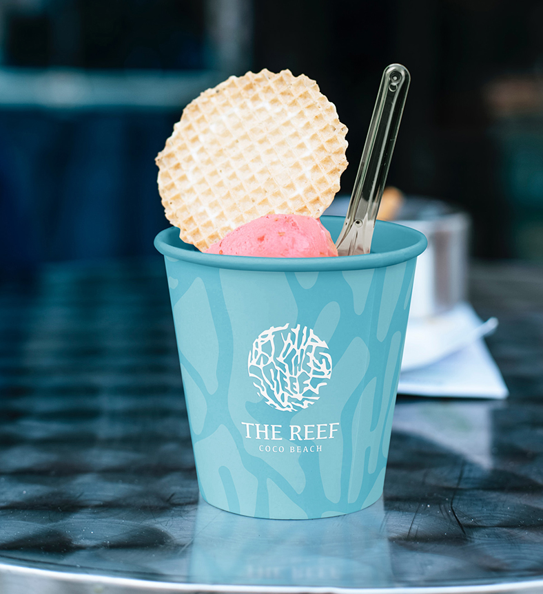 a cup of ice cream with a waffle and spoon, the cup has The Reef logo & branding