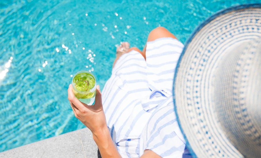 Overhead view of a woman wearing a white hat with her feet in a pool, holding a cocktail at a resort in Grenada.