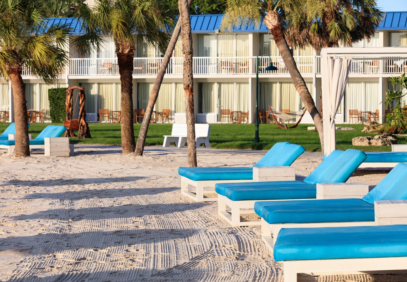 A beach with lounge chairs, palm trees, and a hotel building in the background.