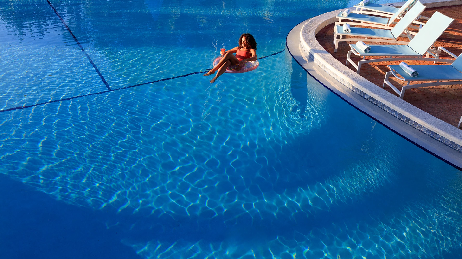 A woman relaxes on a float in a swimming pool with lounge chairs nearby.
