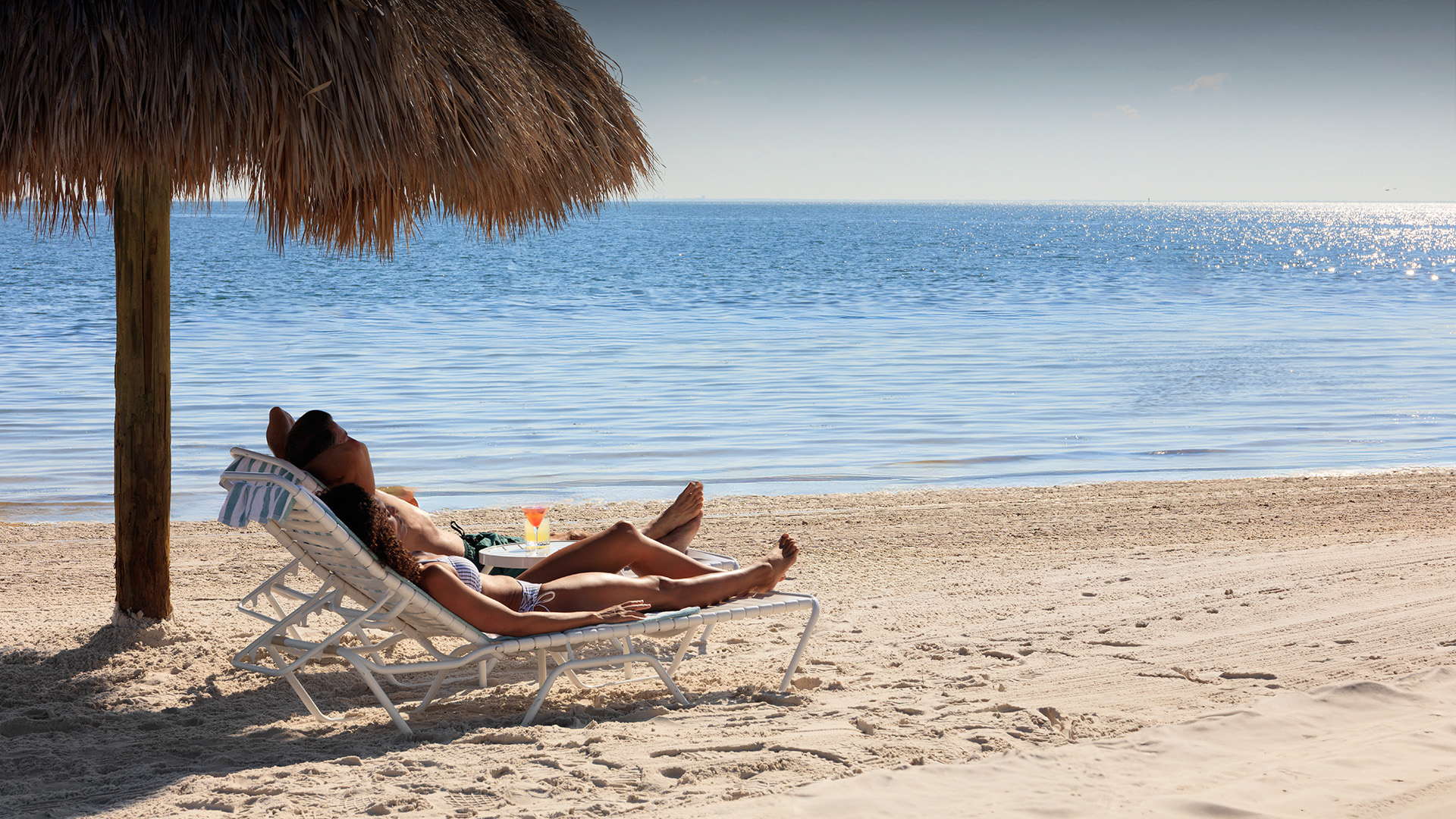 a couple lying on a beach chair under a straw umbrella