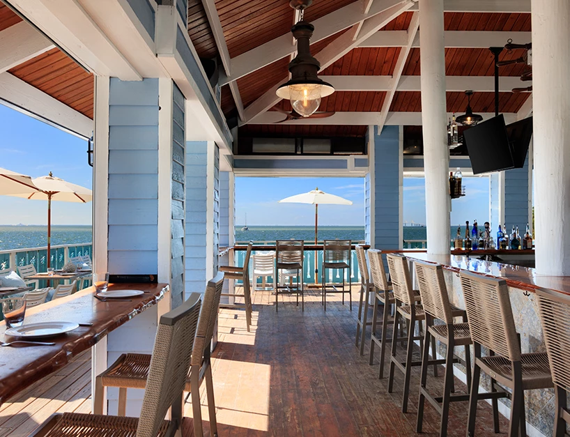 A waterfront bar with stools, a beverage rack, and a TV, overlooking the ocean.