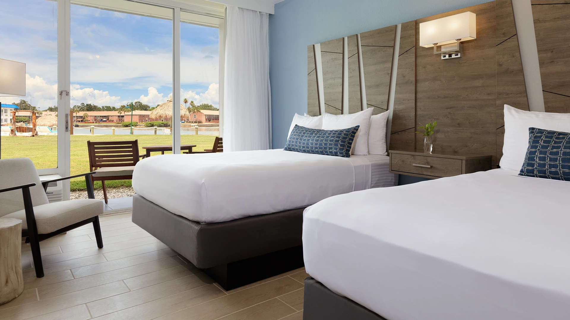 A hotel room with two twin beds, a wooden headboard, a nightstand, an armchair, and a large window overlooking a grassy area.