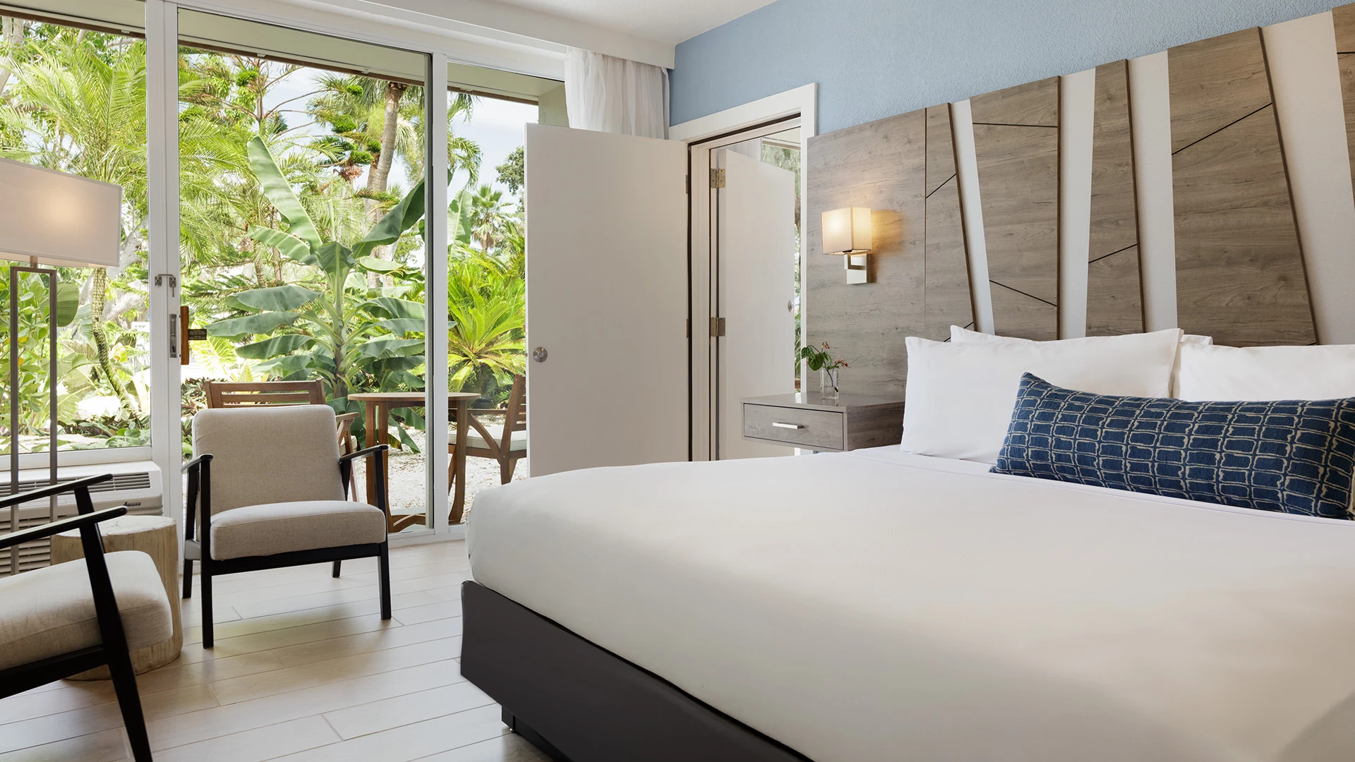 A hotel room with a bed, chair, and balcony door open to a tropical view.