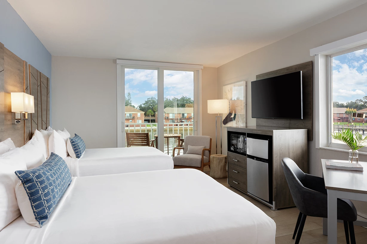 A hotel room with two double beds, a flat screen TV, a kitchenette, and a dining table with a balcony in the background.