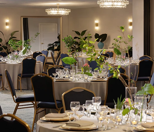 A formal dining room with round tables set for a meal, featuring white tablecloths, glassware, and floral arrangements under elegant chandeliers.