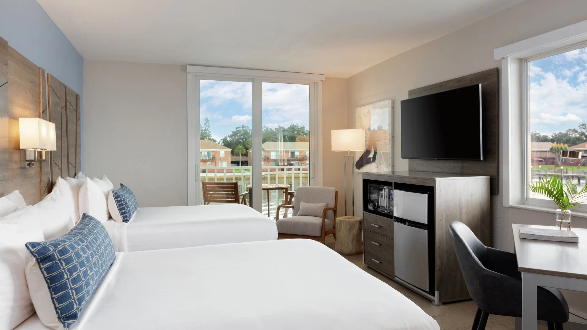 A hotel room with two beds, a desk, TV, mini-fridge, and balcony