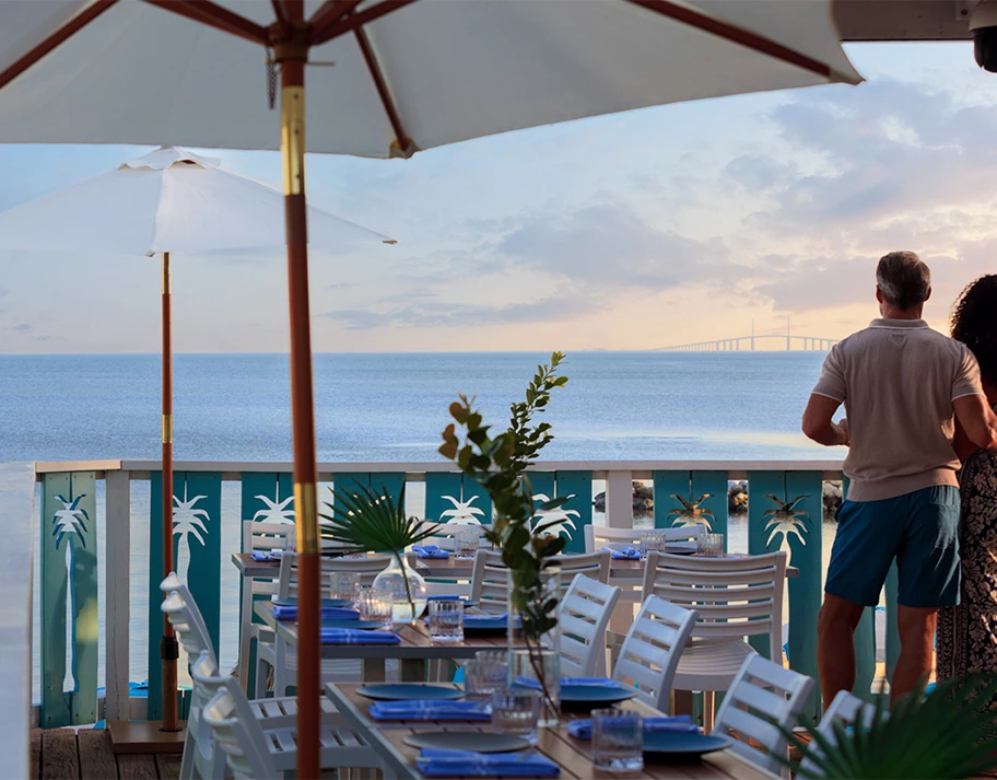 A waterfront dining area with a table set for dinner, an umbrella, and a view of the ocean and a bridge in the distance.