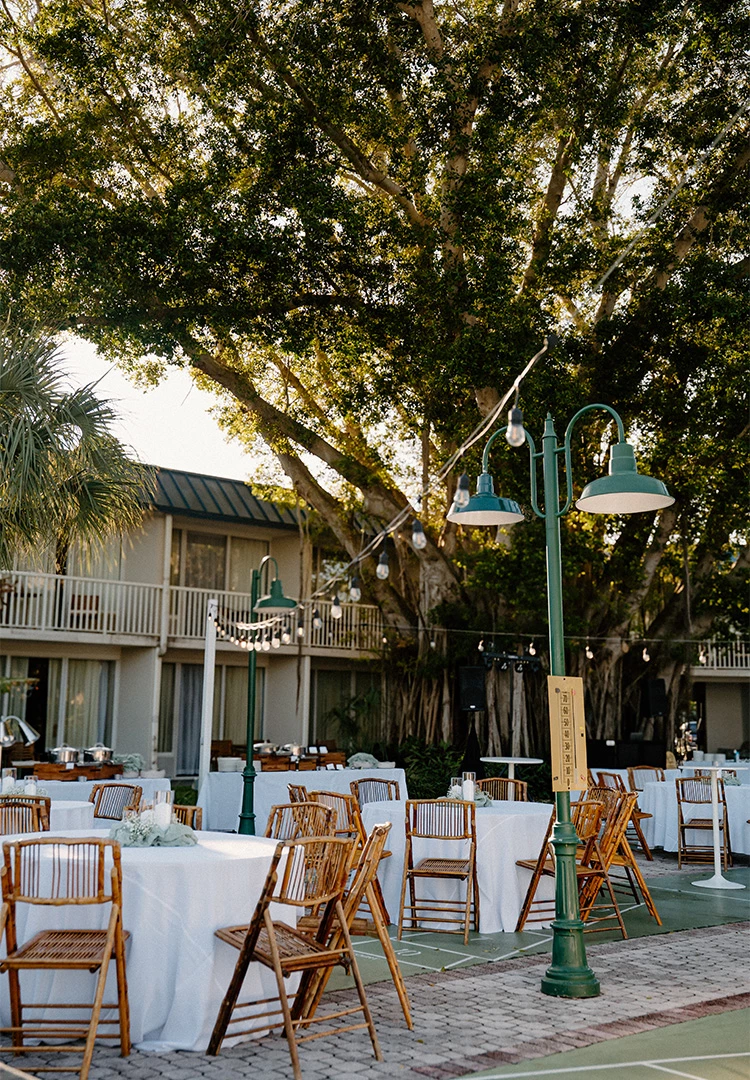 An outdoor event space with round tables, string lights, and a building in the background.