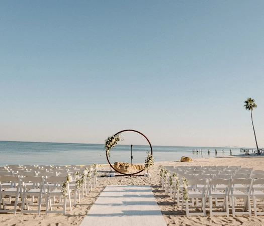 A beach setup for a wedding ceremony with white chairs, a floral arch, and a palm tree in the background.