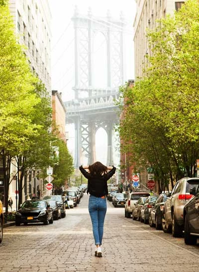 A woman walking down a street with cars and trees on either side, with a bridge in the background.