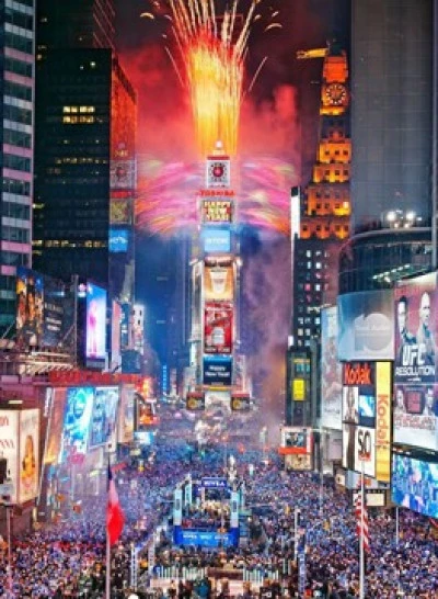 A crowded city street at night with fireworks exploding above a large digital billboard.