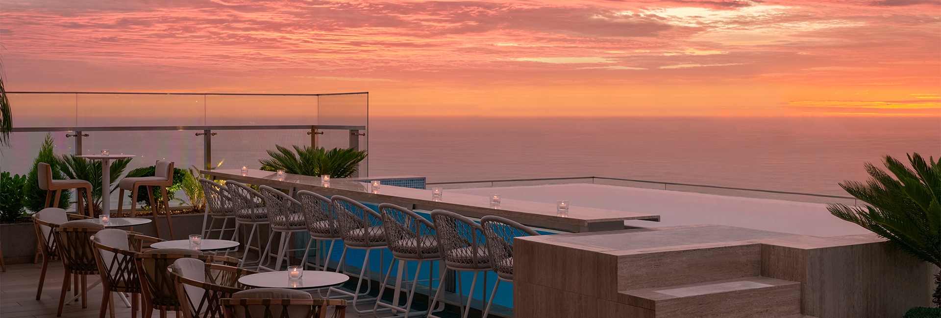 a rooftop bar with chairs and a pool with a sunset in the background
